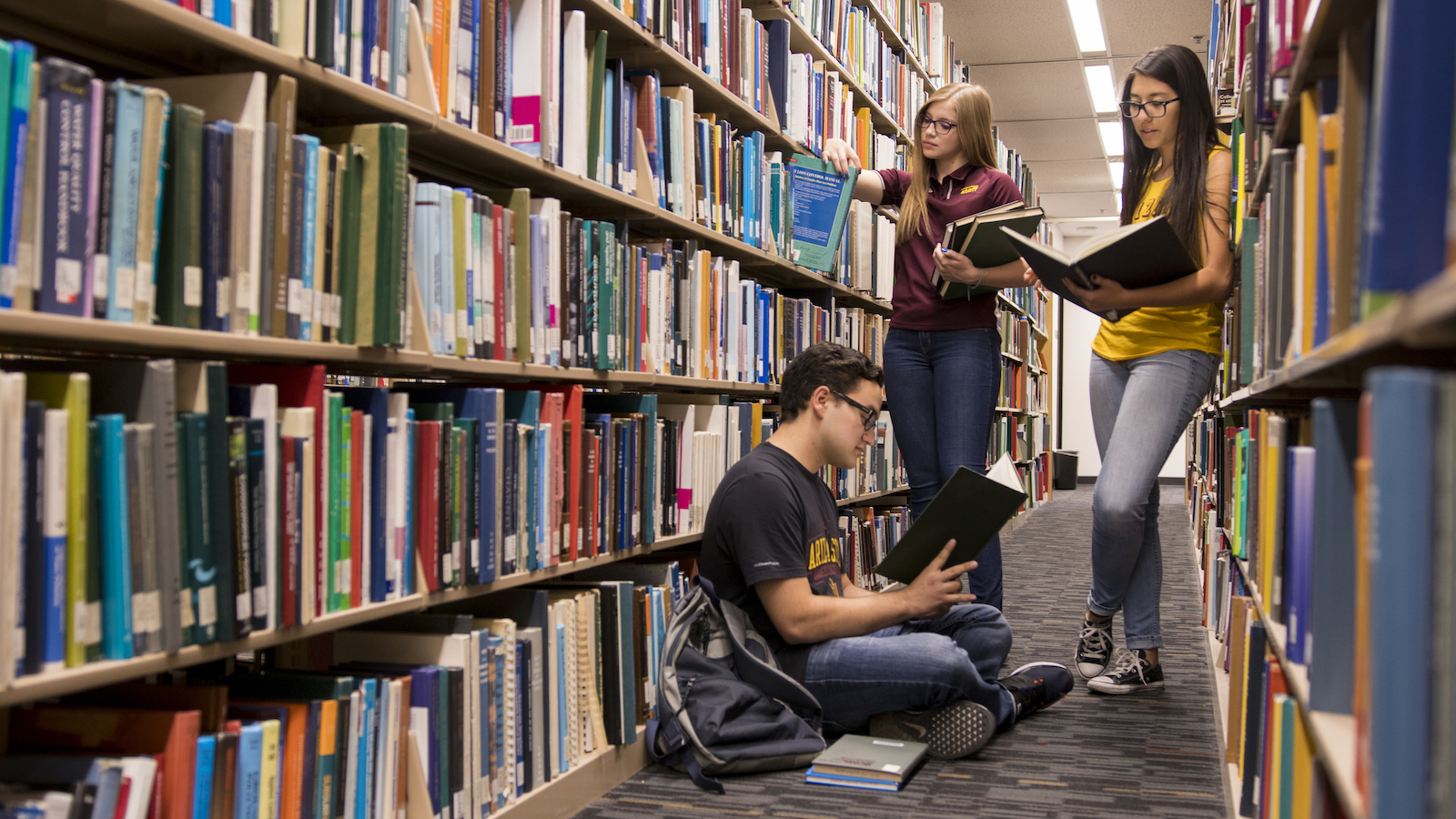 Photo of three students in a library between two bookcases with a caption of "Through reading, you are exposed to new things, new information, new ways to solve a problem and new ways to achieve your goals. Broaden your horizons by checking out a book recommended by Fulton Schools faculty and staff . Photographer Marco-Alexis Chaira/ASU"