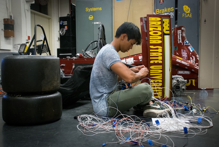 Photo of student sitting on ground working on wiring of car. caption is "Engine crew member Gavin Charoenmin is pictured working on the wiring harness for ASU Formula SAE team’s race cars. Vehicle undergo extensive technical inspections as part of the international Formula SAE competition. Photographer: Alexander Nie/ASU"
