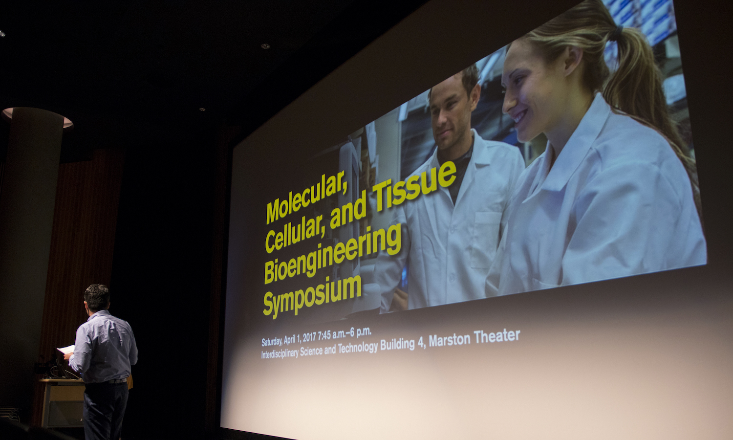 A projection on a screen that says "Molecular, Cellular and Tissue Bioengineering Symposium" over a photo of two people in lab coats.