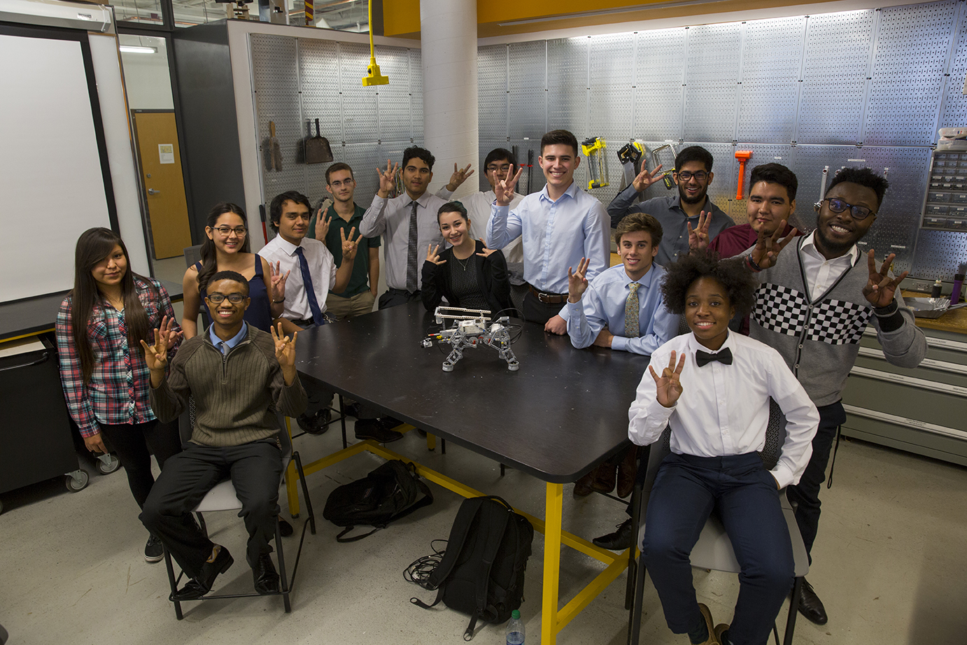 A group of ethnically diverse students gather around a table, displaying the "pitchfork," Arizona State University's signature hand gesture. On the table sits a LEGO robot, and the students are surrounded by tools and equipment. The caption reads: Above: A group of first-generation students pose for a photo during their FSE-100 class. Enrollment of first generation-students has steadily increased in recent years, but retainment remains an issue, with first-gen students 20 percent less likely to complete their degrees in engineering compared to students whose parents attained degrees. But thanks to a recent investment by the National Science Foundation, Arizona State University has embarked on an ambitious project to develop mechanisms, systems and programs to increase persistence of these students as well as cast a wider net to attract underrepresented groups to engineering and STEM careers. Photo by Jessica Hochreiter/ASU  