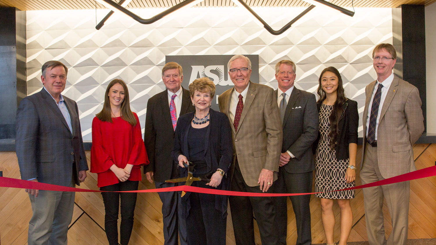 A group of people stand in front of a ceremonial ribbon, woman in center holds a pair of scissors ready to cut.