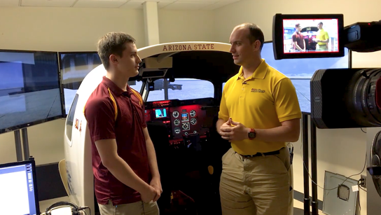 A student and faculty member talk on camera in front of a flight simulator. Caption: Senior Lecturer Benjamin Mertz (right) talks with a student on camera as part of the filming of an online lecture. The Fulton Schools are investing in facilities, tools and staff to enhance course materials for online engineering students. Photographer: Andrew Bautista/ASU