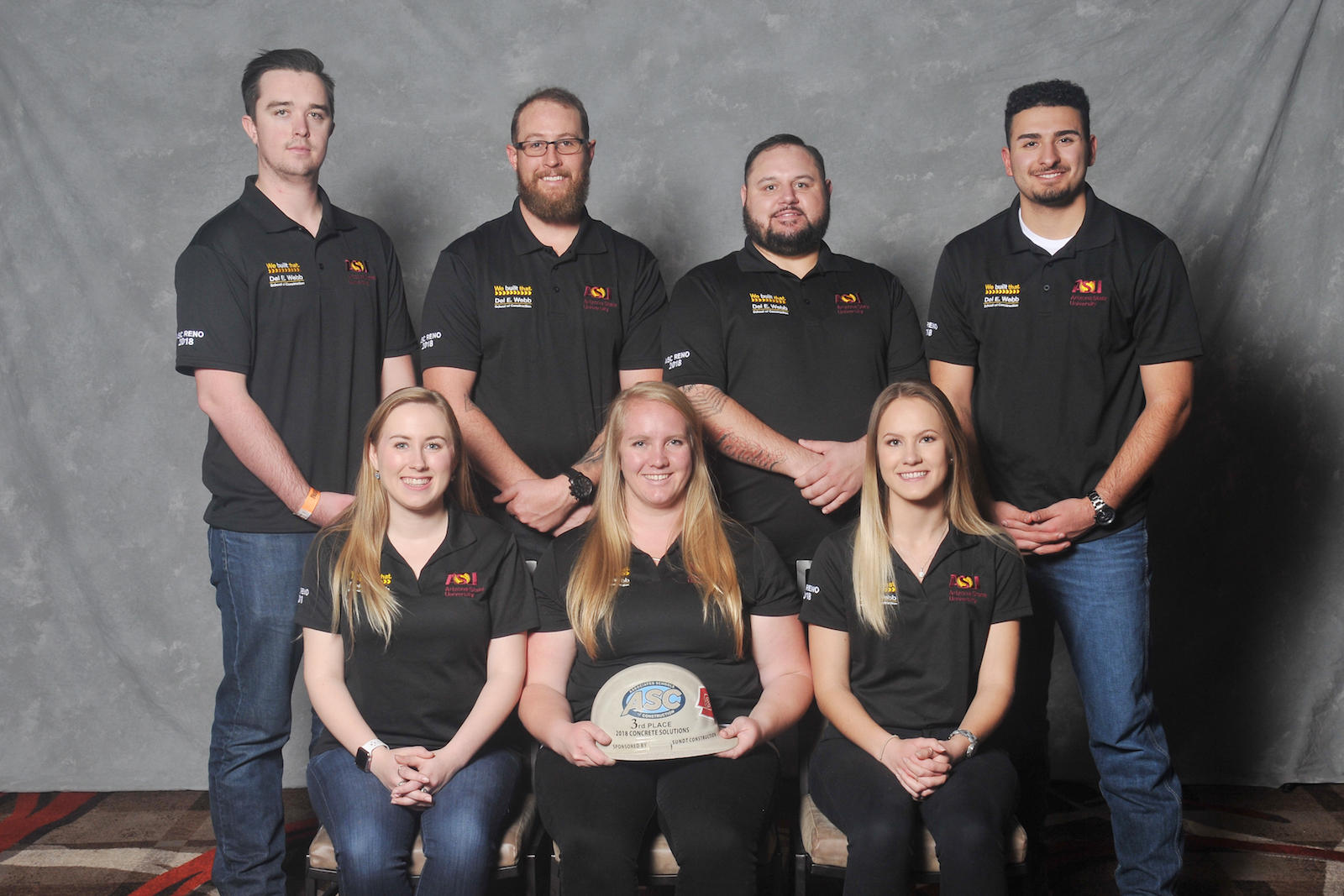 Photo of seven people posing with a hardhat. Caption: Third-place concrete solutions team: (left to right, back to front) Alexander Lee, Jonathan Lyle, Shaun Ford, Corando Chavera III, Hannah Patterson, Kristine Sorensen and Migle Varkalaite. Photo by Associated Schools of Construction for Regions 6 and 7