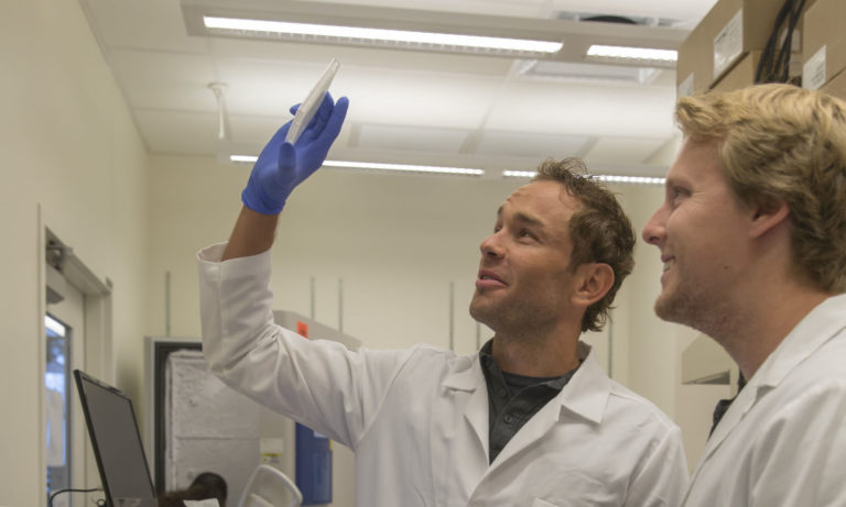 Photo of two men in lab coats with one holding up a dish. Caption: David Brafman (left) and Nick Brookhouser examining a plate used in an assay to characterize the identity of the hiPSC-derived neurons used in their research. Photographer: Jessica Hochreiter/ASU
