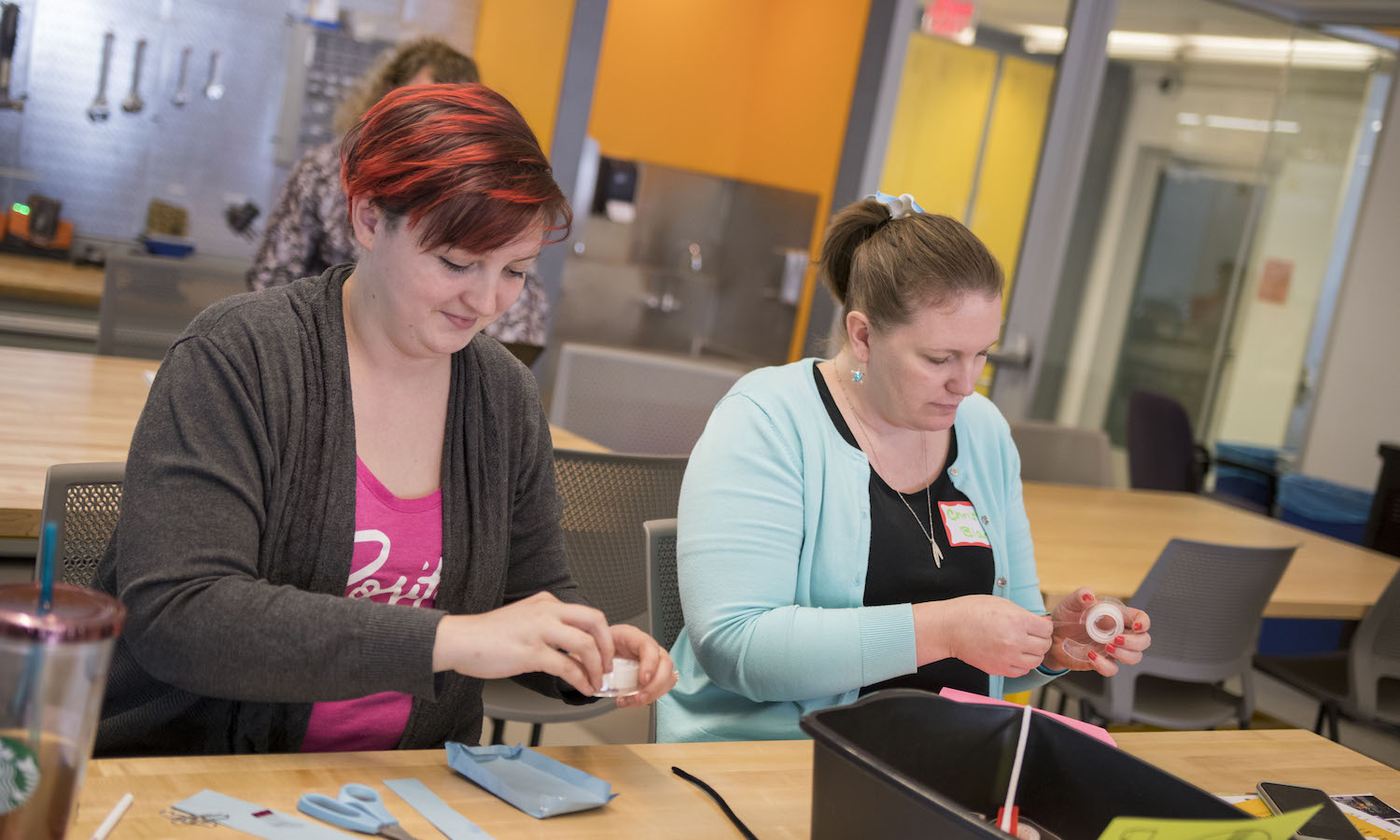 Photo of two women standing and working at a work table. Caption: