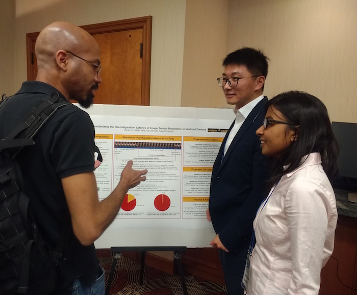 Photo of student presenting to two others in front of a poster. Caption: Computer engineering doctoral student Jinhan Hu (second from right) discusses his research on reducing mobile vision application power consumption with other researchers attending ACM HotMobile ‘18. Hu presented one of four demonstrations — his first public talk at a conference. Photo courtesy of Robert LiKamWa.