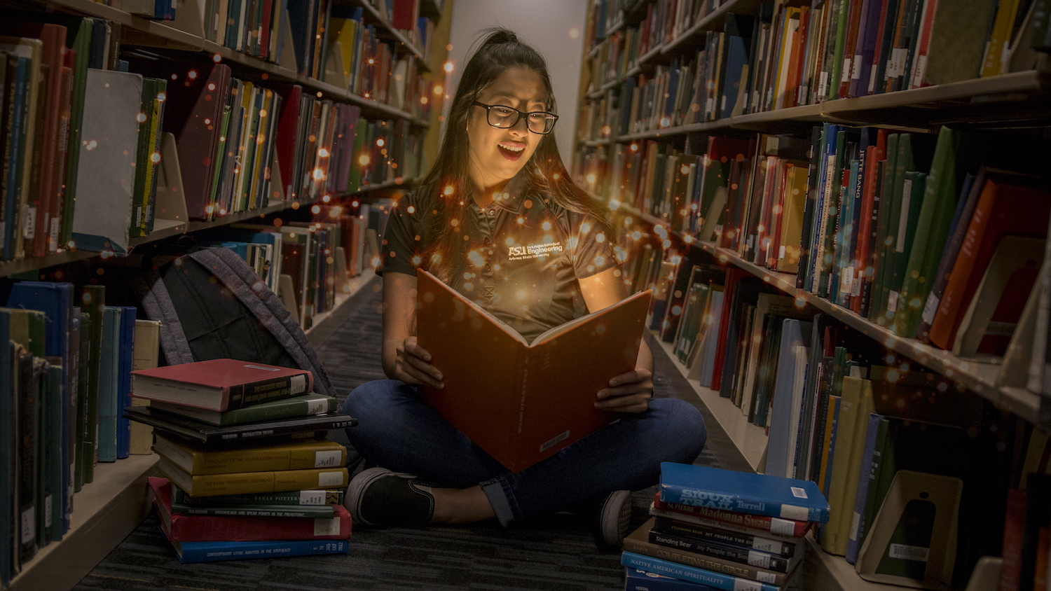 image of a girl sitting on the floor of a library surrounded by books with light particles glowing from a book she is holding