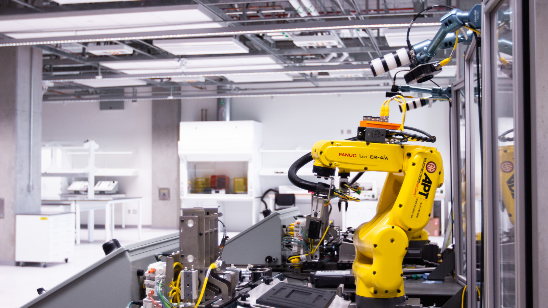 A robotics instructional lab with a FANUC ER-4iA robotic arm fitted with a gripper and camera system. The workspace includes automated assembly machinery and surrounding workbenches in a clean, industrial environment.