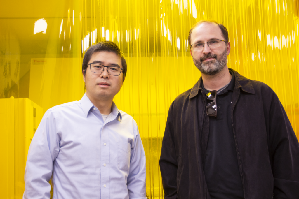 Two men stand side by side inside the Advanced Manufacturing and Functional Device Laboratory, in front of a bright yellow UV light seal curtain.