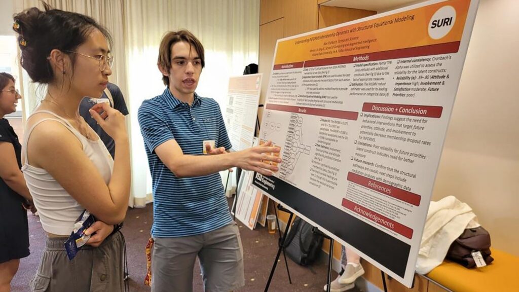 Alec Fishbach points at a poster at a research symposium while a guest looks on.