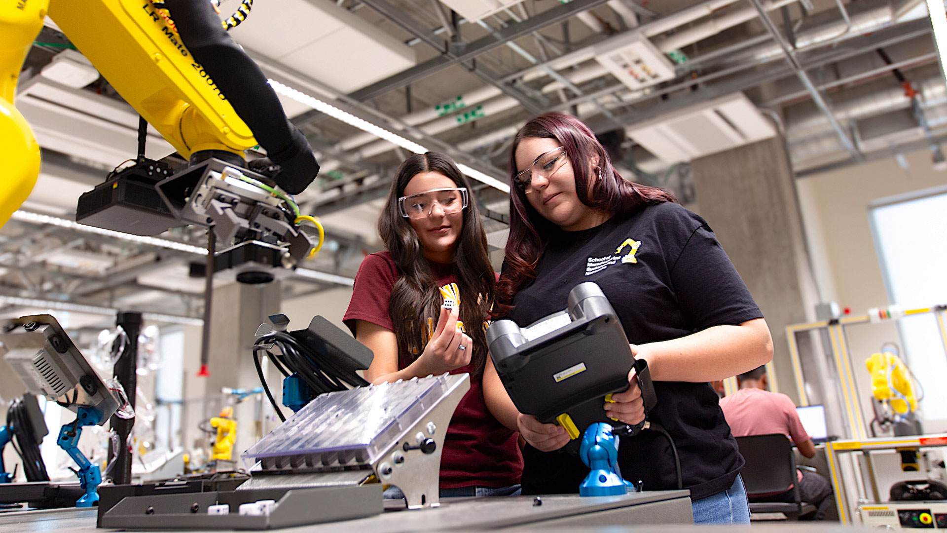 Jenavieve Echegaray and Lexana Echegaray wear safety glasses while working beside a yellow FANUC industrial robotic arm in the Robotics Systems Instructional Lab located in ISTB 12.