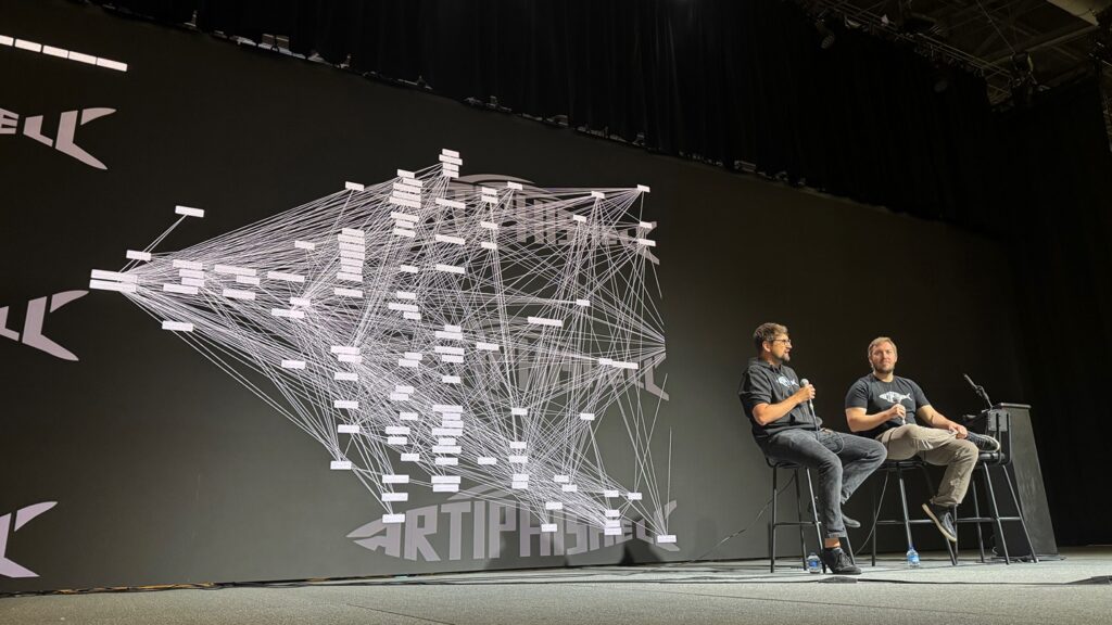 Gibbs and Lukas Dresel sit in chairs on stage in front of a background showing a technical illustration of computer code.